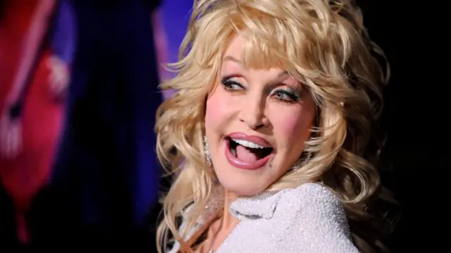 Dolly Parton smiles as she stares to her right. She is wearing a silver glittery outfit