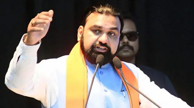 Samrat Choudhary is pictured speaking behind two microphones, gesturing with his right hand.