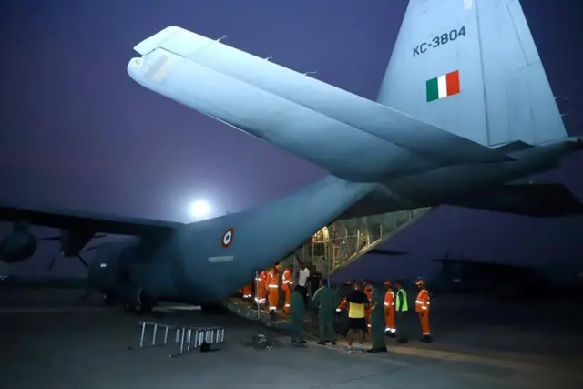 workers load aid onto a plan destined for nepal. they wear orange high-vis suits. the plane appears to be a military aircraft marked with the air force logo.