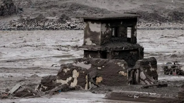 The ruins of a building are shown covered in mud and debris beside a swollen river.