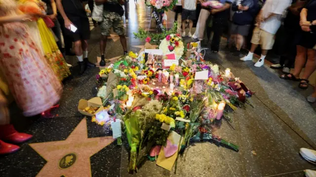 Flowers laid across a pink Hollywood Walk of Fame star as people stand around it in a circle