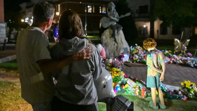 A couple stand together in the foreground in front of a Dolly Parton statue. A young child is stood in front of them and is looking down at some flowers placed near the statue
