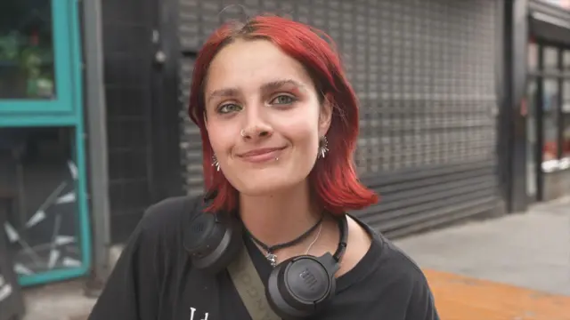 Person with short red hair in the foreground, wearing a black T-shirt, black over-ear headphones around the neck, a cross-body strap, a nose ring and earrings. The person is facing the camera on a pavement outside a building. Behind the person is a dark tiled wall and metal security shutter, with a turquoise-framed doorway visible on the left and part of a street scene on the right. No bags, bins or markers are visible.