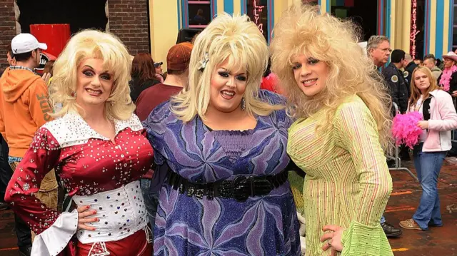 Three men dressed in colourful Dolly Parton drag outfits smile at the camera