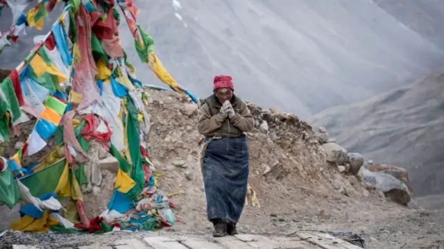 A person is shown walking up a mountain beside strings of colourful prayer flags.