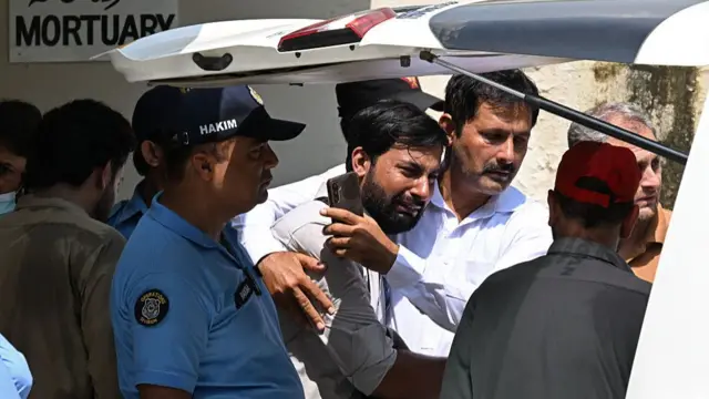 A man looks distraught and is held by another man next to him just after a body has been put in the back of a car. A sign on the wall behind reads "mortuary"