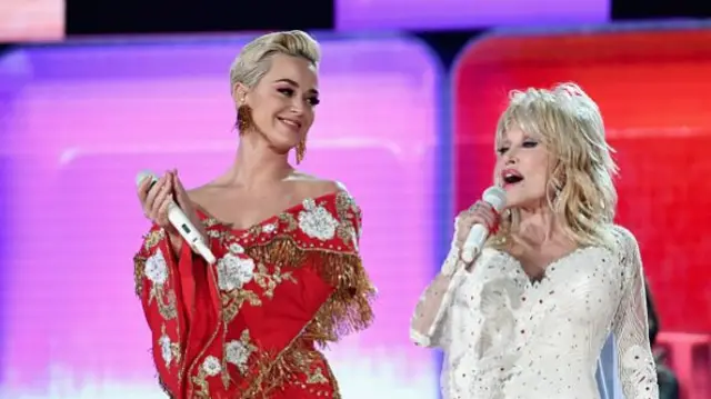 Dolly Parton holds up a microphone as she sings as Katy Perry smiles and looks on