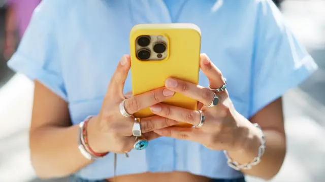 A woman wearing rings and bracelet holds a yellow phone.