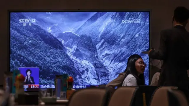 A woman sits in front of a news screen talking to a man whose back is to the canmera. The screen broadcasts news footage of a flash flood and mudslide on the border of the Tibet and Nepal