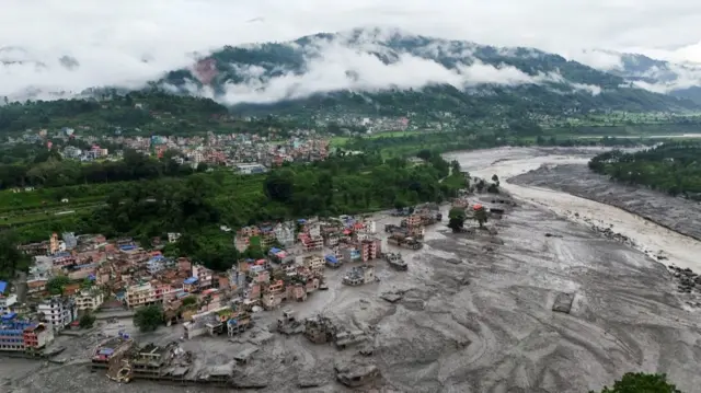A drone shot shows several houses on the bank of the Trishuli river. In the foreground, the houses and roads closest to the river bank are covered in mud, while in the background more groups of house can be seen in the distance seemingly unaffected by the floods