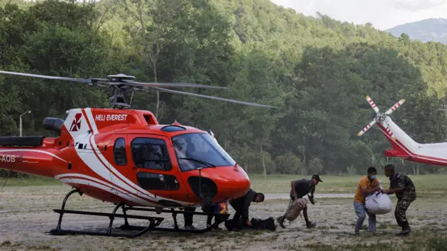 A red helicopter with a white stripe sits in the middle of a muddy area with trees in the background at Maithali Barrack, an army training centre, which serves as a primary base to airlift survivors following flash floods and mudslide.