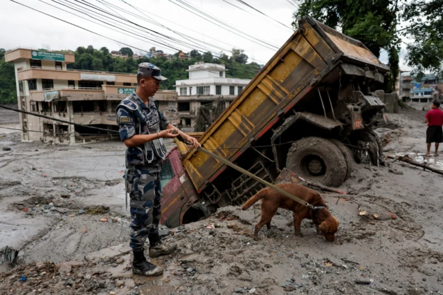 A member of the Nepalese security forces follows a sniffer dog through a mud-covered area of Trishuli in Nuwakot district. A partially tilted truck faces downwards as its rear wheels grip part of collapsed pavement