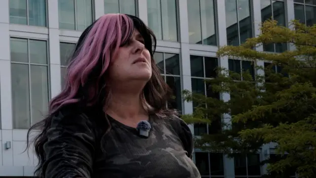 A woman with dark hair with a large pink streak stands outside a courthouse