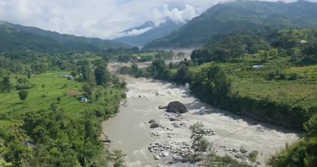 A screenshot of a video of the Trishuli River, there are fields and buildings on either side of it and the floodwater is beginning to rush down the river.