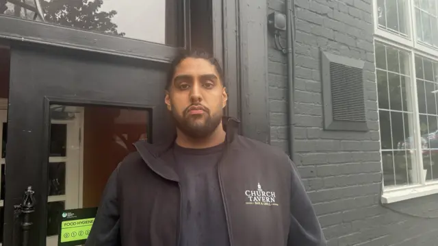 A man stood outside a pub wearing a black top and black gilet
