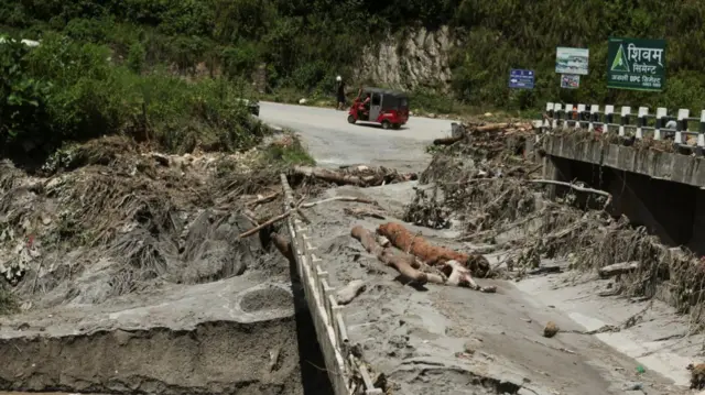 A road in Nepal with a partially collapsed bridge covered in debris, including tree branches, and mud