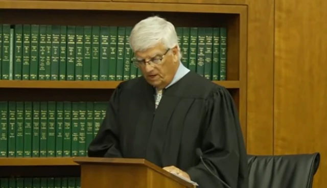 A judge in black robes stands at a lectern in front of a bookcase filled with green bound books