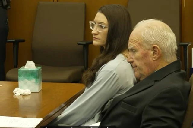 A woman with long dark hair and glasses sits beside an older man in a suit with a box of tissues on a table in front of them