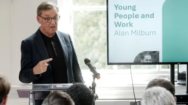 Milburn speaking at a lecturn in front of a screen which says 'young people and work, alan milburn'