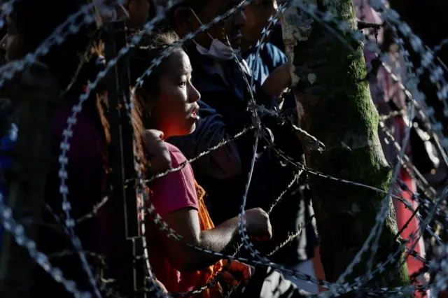 A young girl looks to the right while standing on next to a barbed wire fence. The girl is seen in the photo encircled by rings of barbed, which appear in the foreground