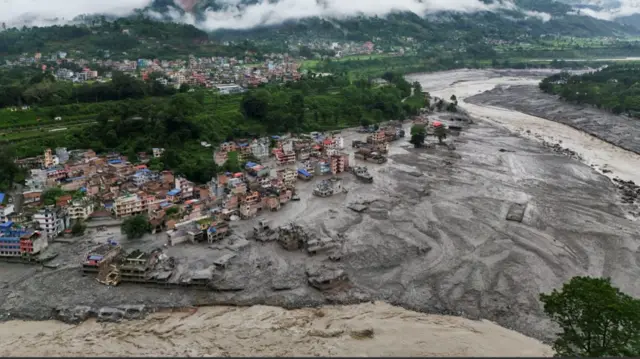 Wide shot of a village partially submerged by mud following flooding in the Nuwakot district of nepal. Houses are partially submerged as a muddy stream rushes by on the right side of the frame