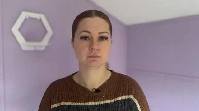 A close-up of a woman with tied back brown hair, wearing a dark brown jumper. The purple walls of her cabin are behind her.