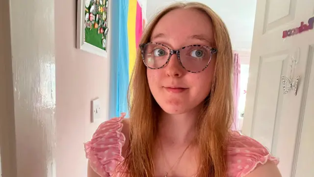 Rebecca looking at the camera smiling. She's wearing glasses, wearing a pink dress