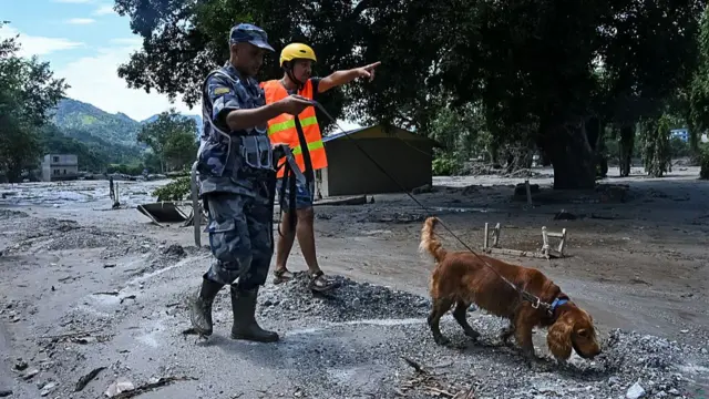 A man clad in grey camouflage military uniform holds a spaniel-type looking dog on a lead, while wading through mud and debris. He is standing next to another man in a hi-vis jacket and helmet who is pointing towards something ahead of them