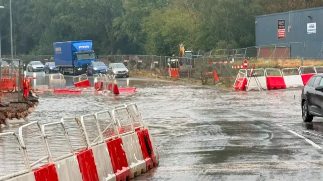 A queue of cars lined up on the far side on a road, which has flooded in front of them. There are red and white roadworks barriers either side of the road.