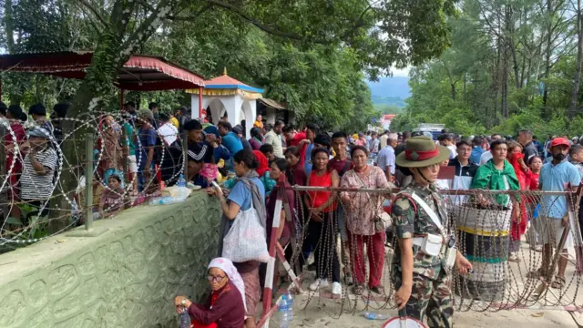A large crowd of people is queueing outside a military facility beside a road surrounded by trees. The crowd is standing next to a wall which has barbed wire on top, while a military official stands guard