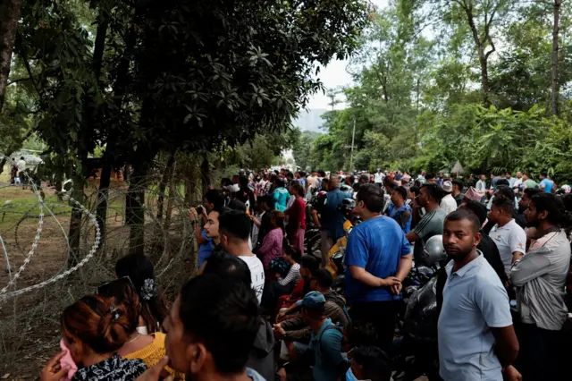 A large group of people stand behind barbed-wire.
