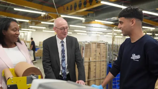 Pat McFadden, an older man in a suit, talking to a young man in a Boots warehouse.