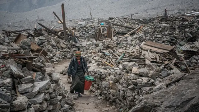 A woman carrying a red bucket walks along the remains of a village destroyed by an avalanche. She's walking along the path, debris, stones and wood on both sides