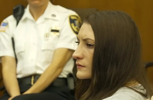 A woman with long dark hair looks on in court as a law enforcement officer stands in the background