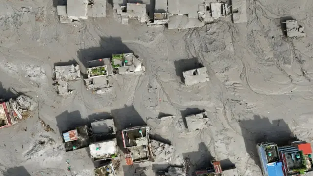 Aerial image showing the aftermath of a large landslide. Focus is on a village, homes buried in mud with only the roofs visible
