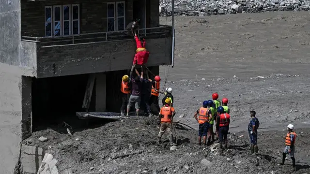 A group of people wearing protective equipment stand on some rubble at the base of a house. The rescuers closest to the house are helping lower a person down from the balcony