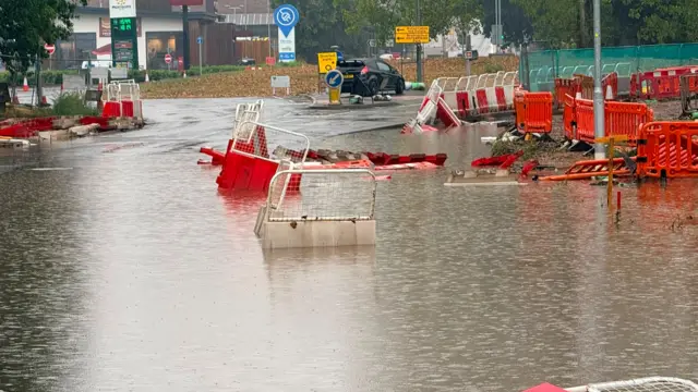 Floodwater covers a section of a road where red‑and‑white construction barriers are partly submerged, some knocked over and floating