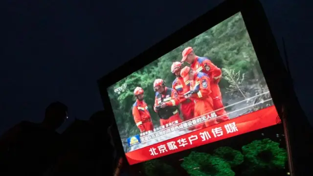 A large screen is shown at night-time, with five rescue workers in orange overalls clustered in an outdoor area in front of vegetation.