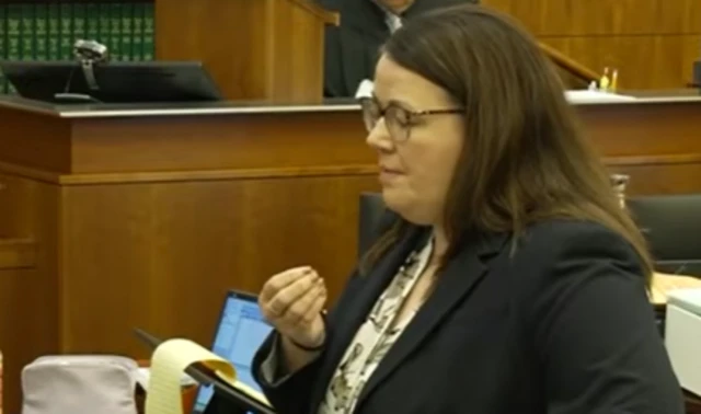 A woman in glasses stands in a courtroom speaking to the jury