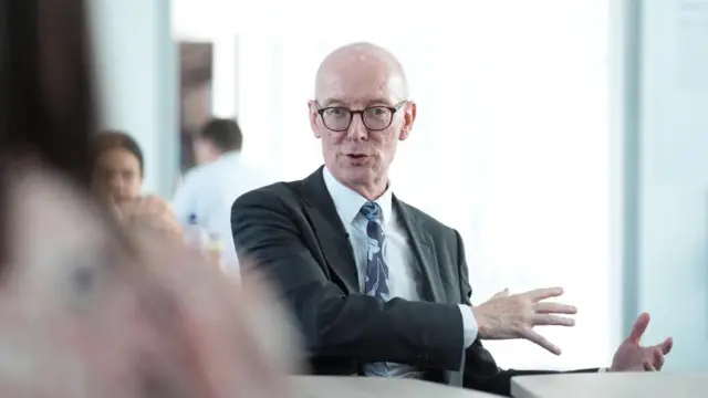 An older man with glasses and suit sitting in a room, talking.
