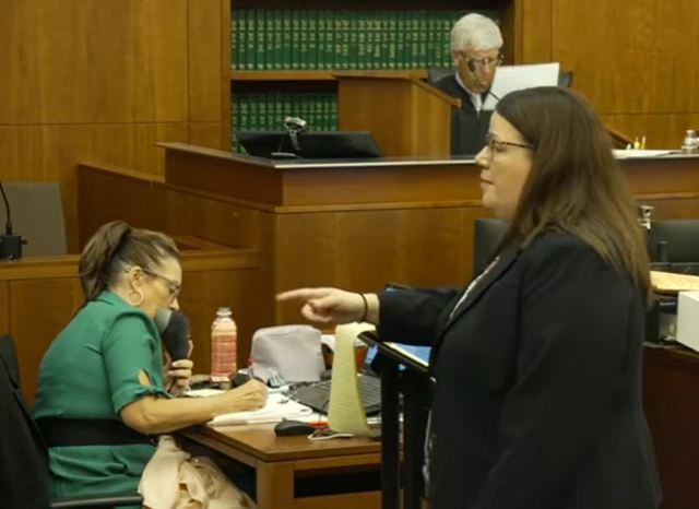 A woman in suit points in a courtroom with a judge and stenographer seated behind her