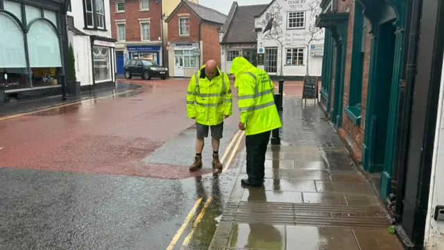Two people wearing hi-vis jackets look down at a drain on the side of a road in a town centre.