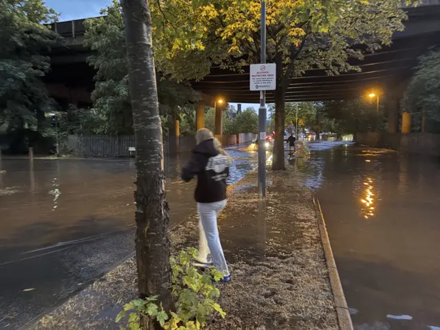 A road completely covered with surface water. A woman is stood on a central bit of pavement