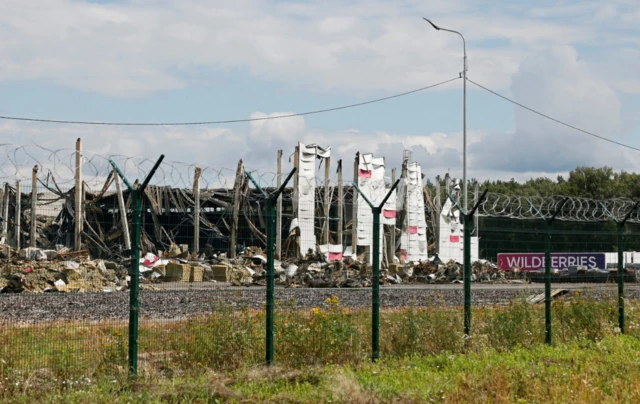 Remains of the internal structure of a Wildberry site in Elektrostal photographed from a field outside the building's perimeter. Parts of the external walls (in white and purple) remains up, a Wildberries' lorry is parked in the background
