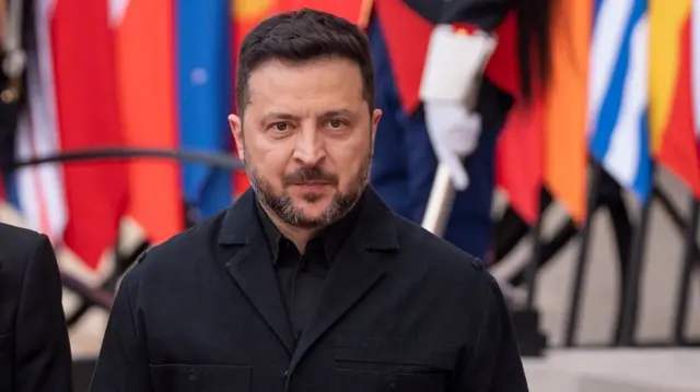 Volodymyr Zelensky is pictured looking solemn wearing a black shirt and coat in front of a row of flags.