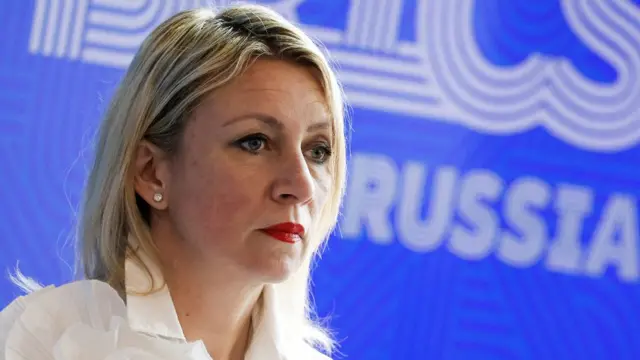 A woman with shoulder-length blonde hair, wearing a white shirt, in a close up shot in front of a blue sign saying "Russia"