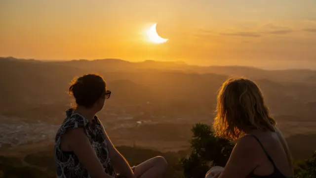 Two women sat on a hill watching a partial solar eclipse in Menorca, Spain