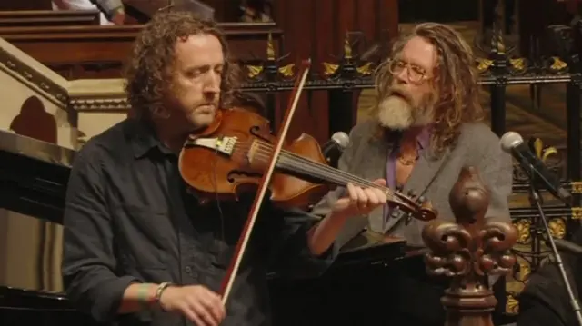 One man wearing a black shirt holds a violin with both hands. He has long brown hair. Another man with long brown hair and a grey beard plays a black piano.