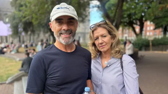 Two people look at the camera. On the left, a man in a navy t-shirt and grey cap, on the right a woman with long blonde hair and a grey shirt. They stand on a red-brick path.