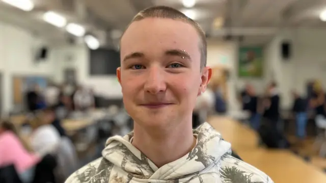 AJ has cropped fair hair and wears a green camouflage hoodie. He smiles and is in a room with people sitting at tables behind him